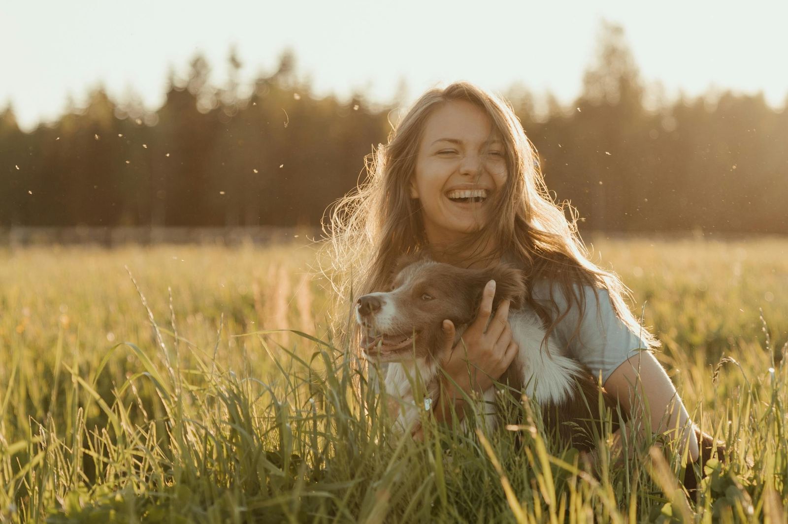 A woman laughing joyfully with her dog in a golden sunlit field