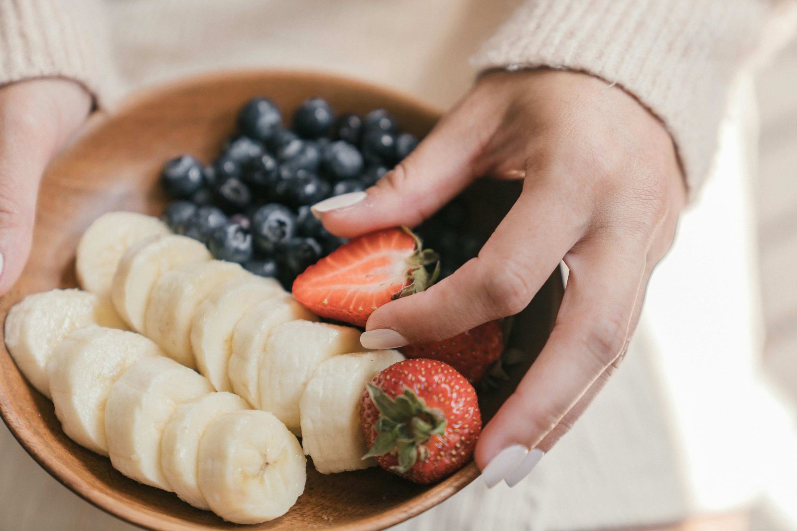 Hands holding a wooden bowl of fresh fruit — banana, blueberries, strawberries — nourishing the body with intention