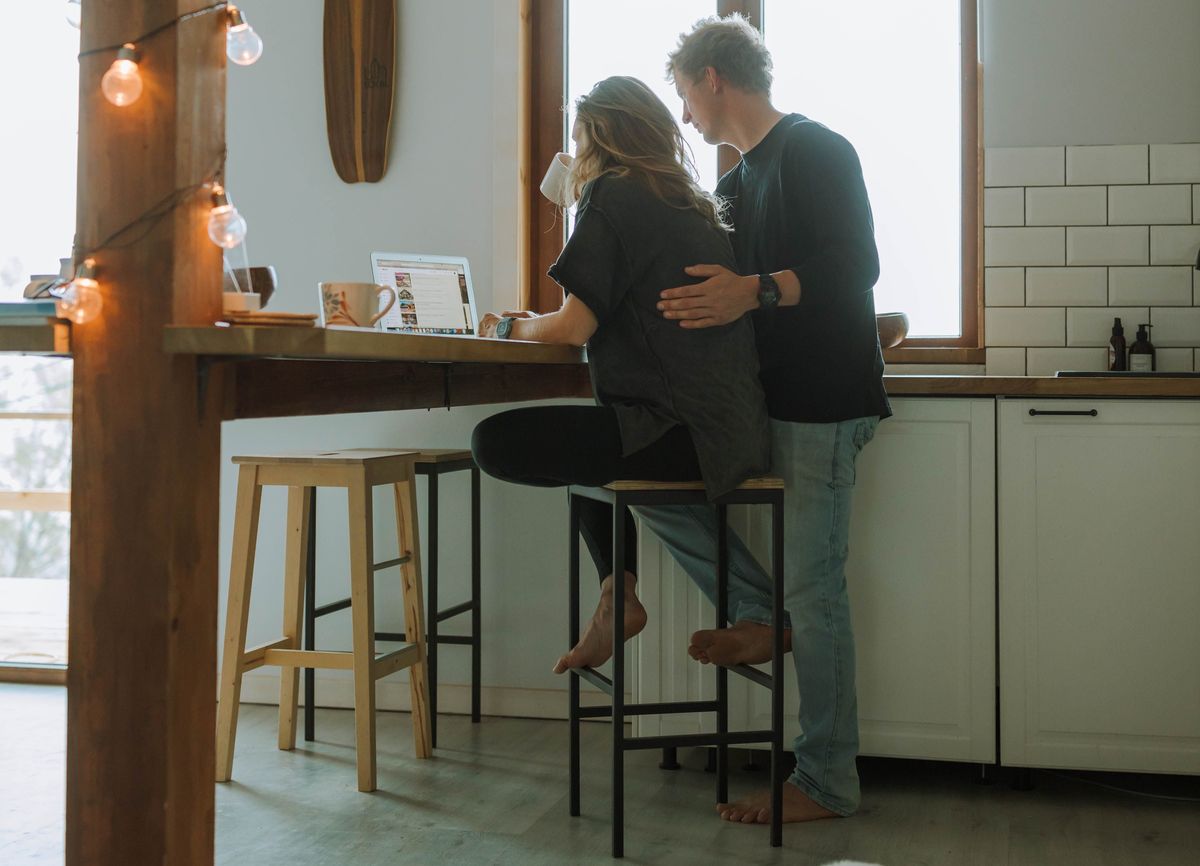 A couple standing quietly together in the kitchen, his arm around her as she rests by a laptop and coffee