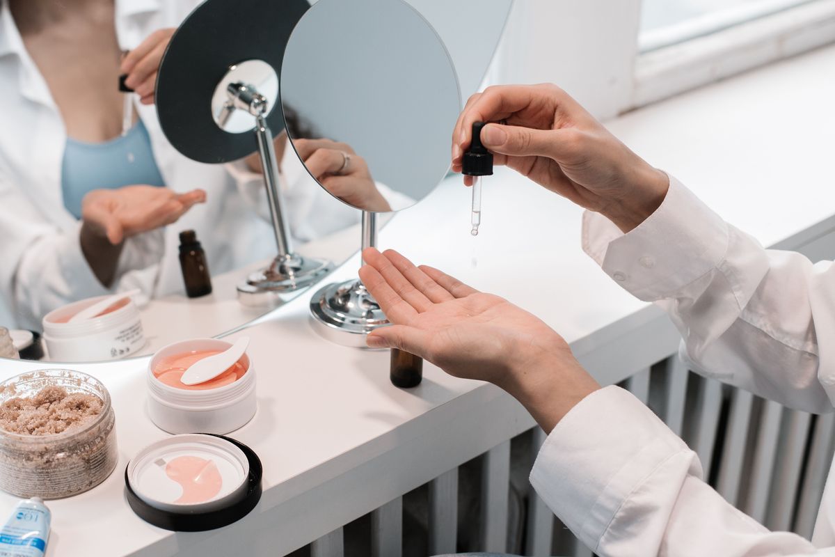 A dropper bottle being used at a mirrored vanity surrounded by skincare jars and serums