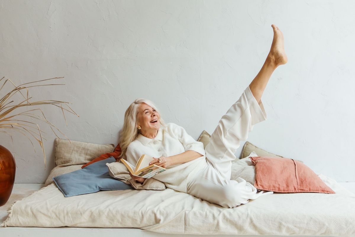 A woman laughing mid-book, kicking a leg up playfully on a daybed — the look of someone genuinely young at heart