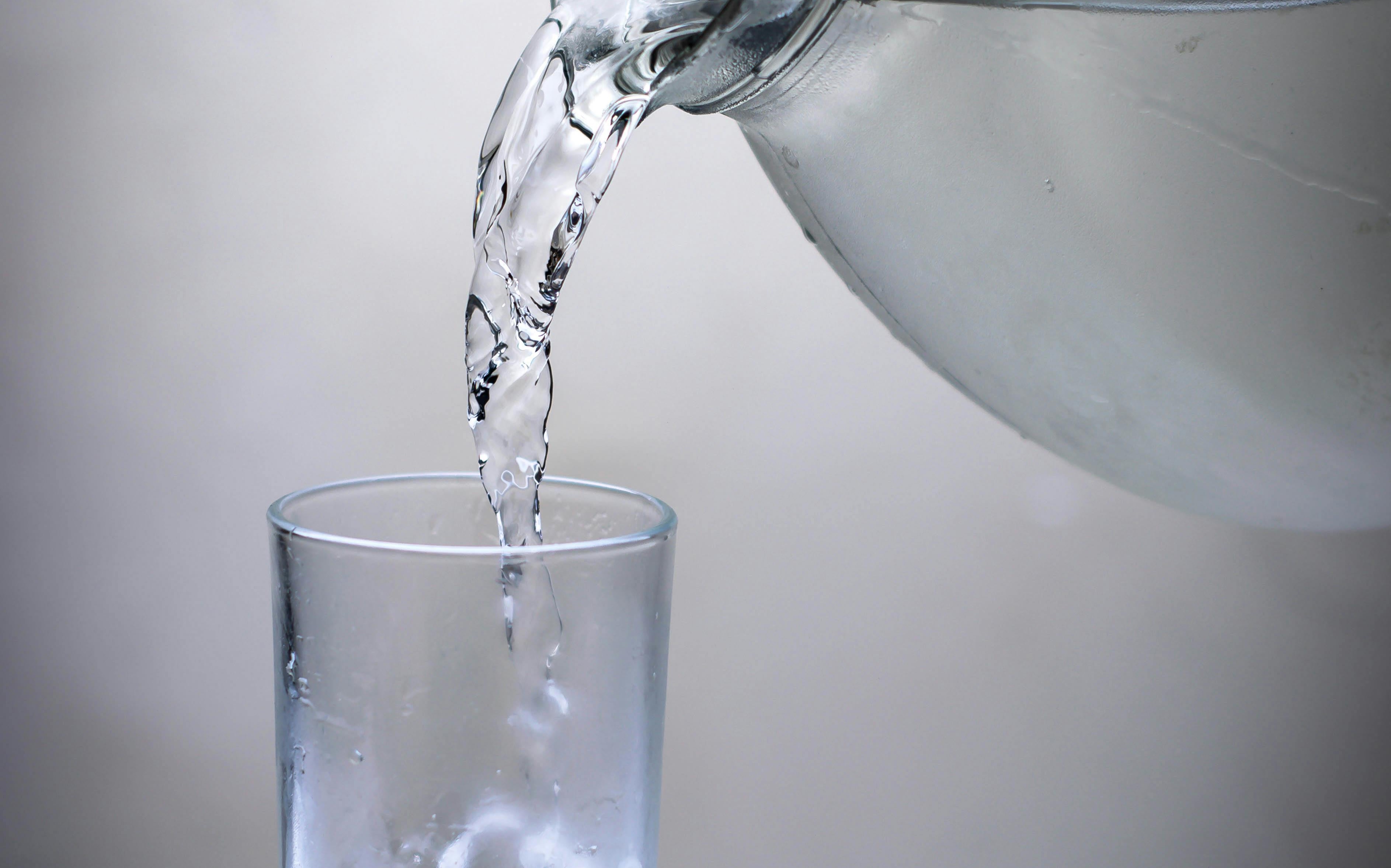 Water pouring from a glass jug into a clear drinking glass against a clean gray background