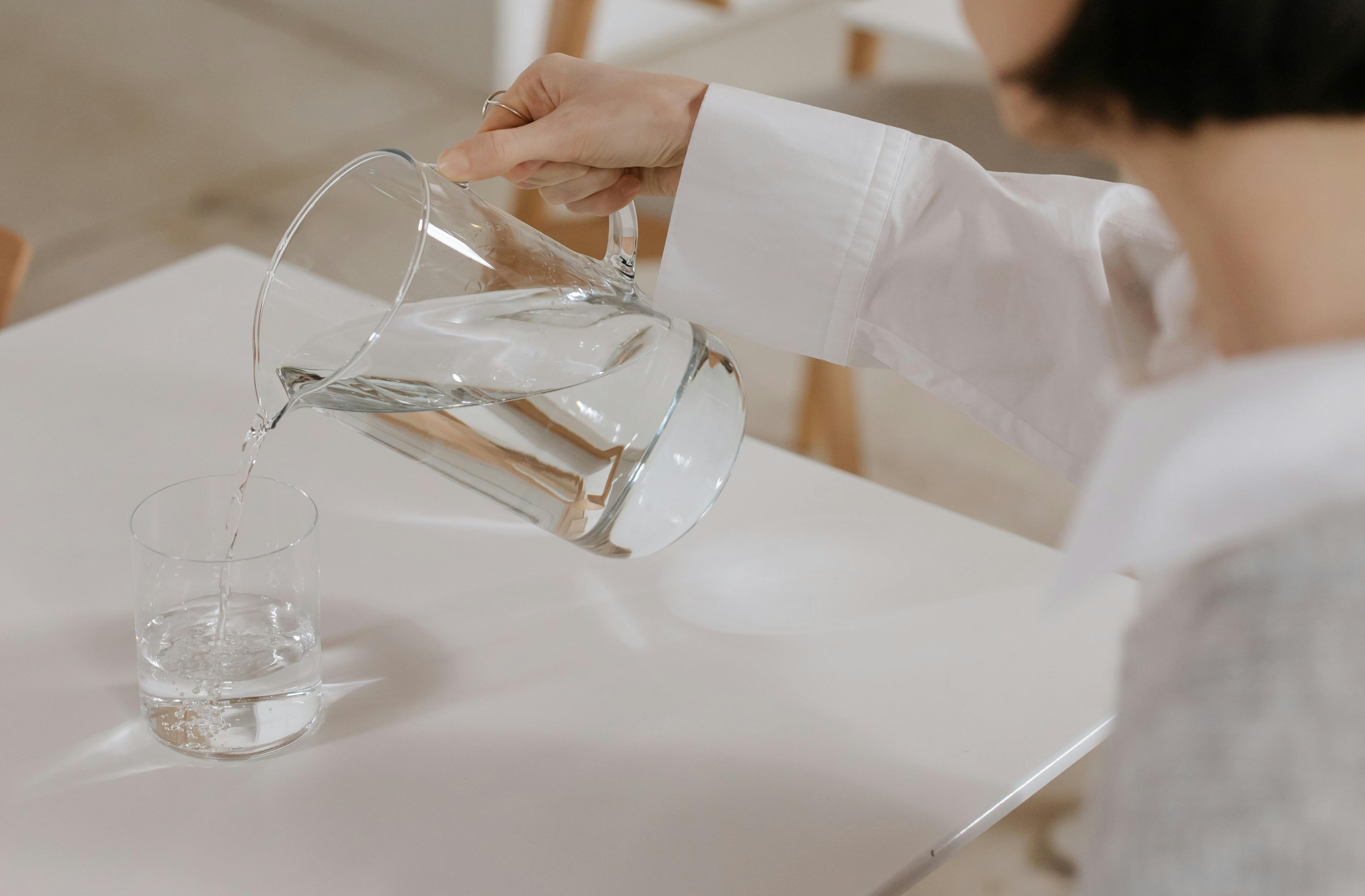 A hand pouring water from a glass pitcher into a small glass on a white table