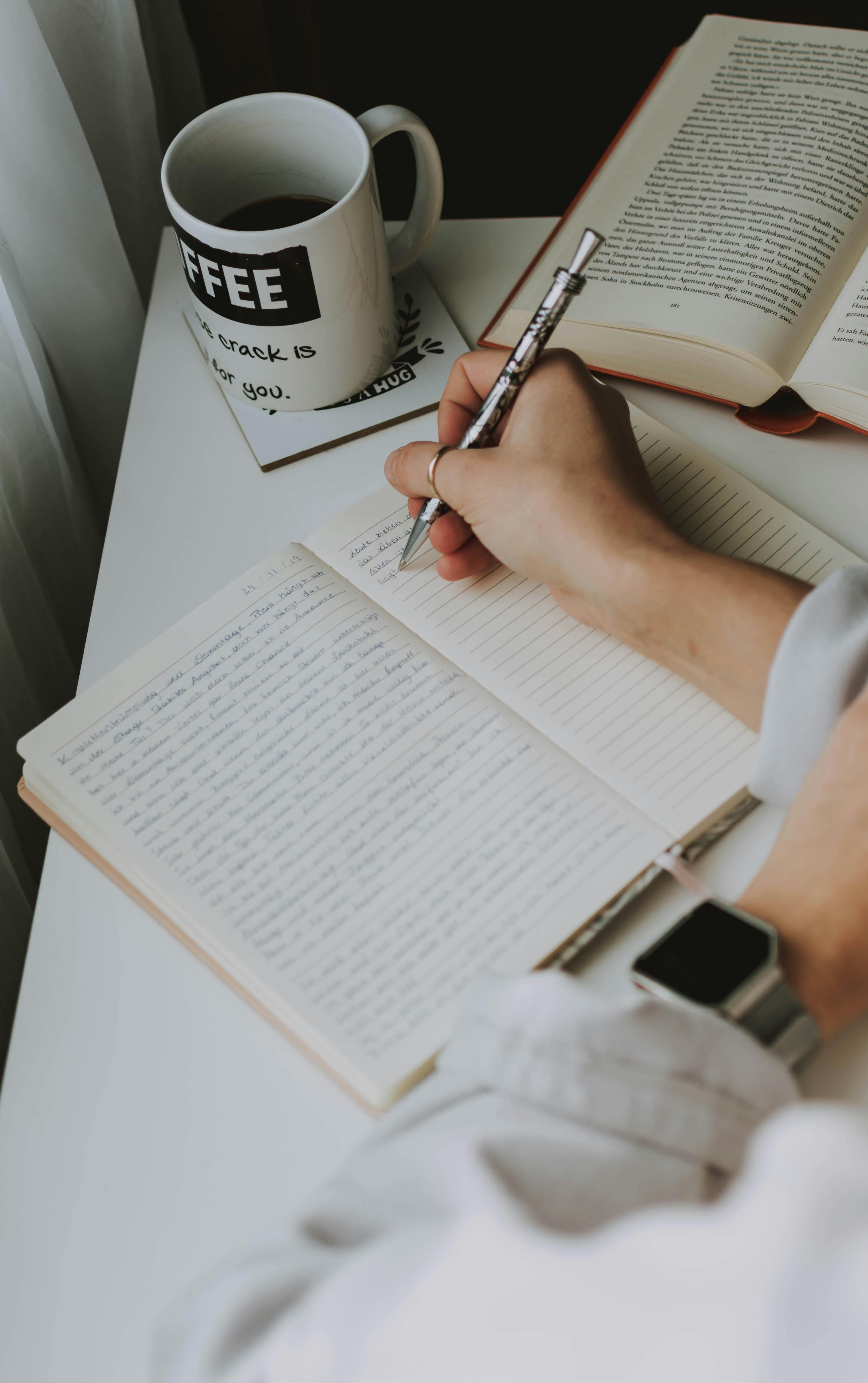 A hand writing longhand in an open notebook beside a coffee mug, soft morning light