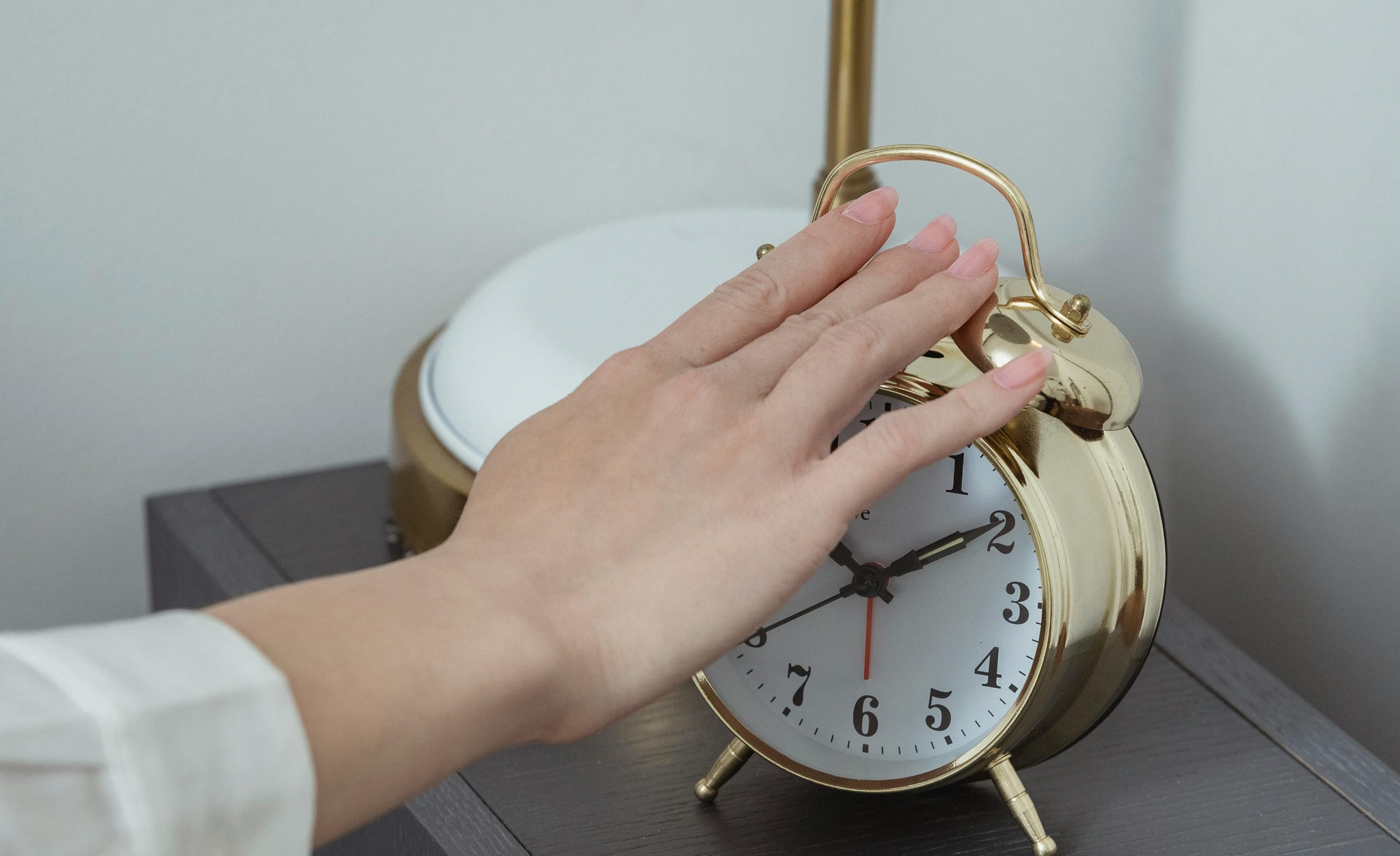 A hand reaching to silence a gold alarm clock on a bedside table, early morning