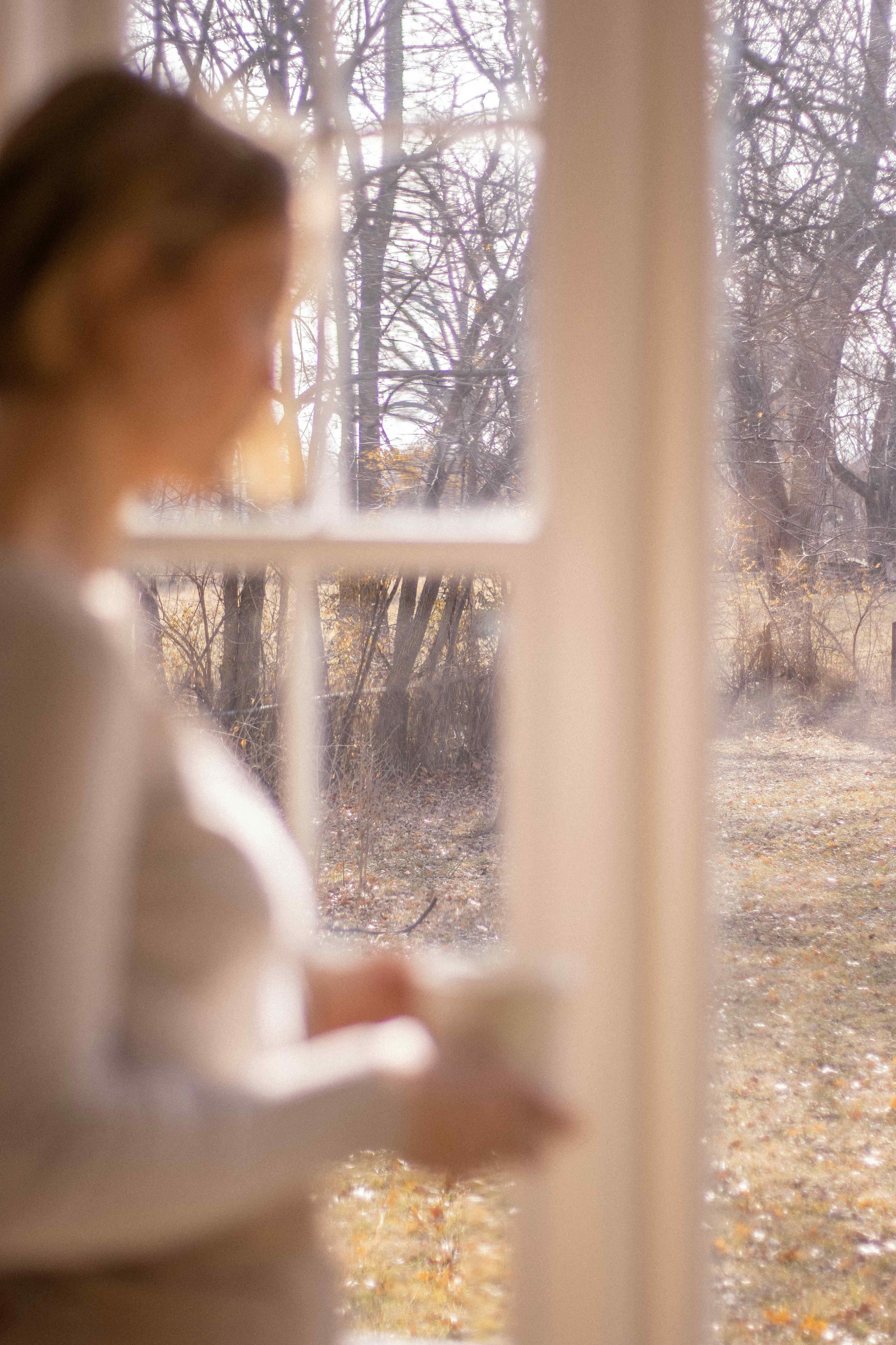 A woman holding a warm mug, looking out a window at bare winter trees, soft and contemplative