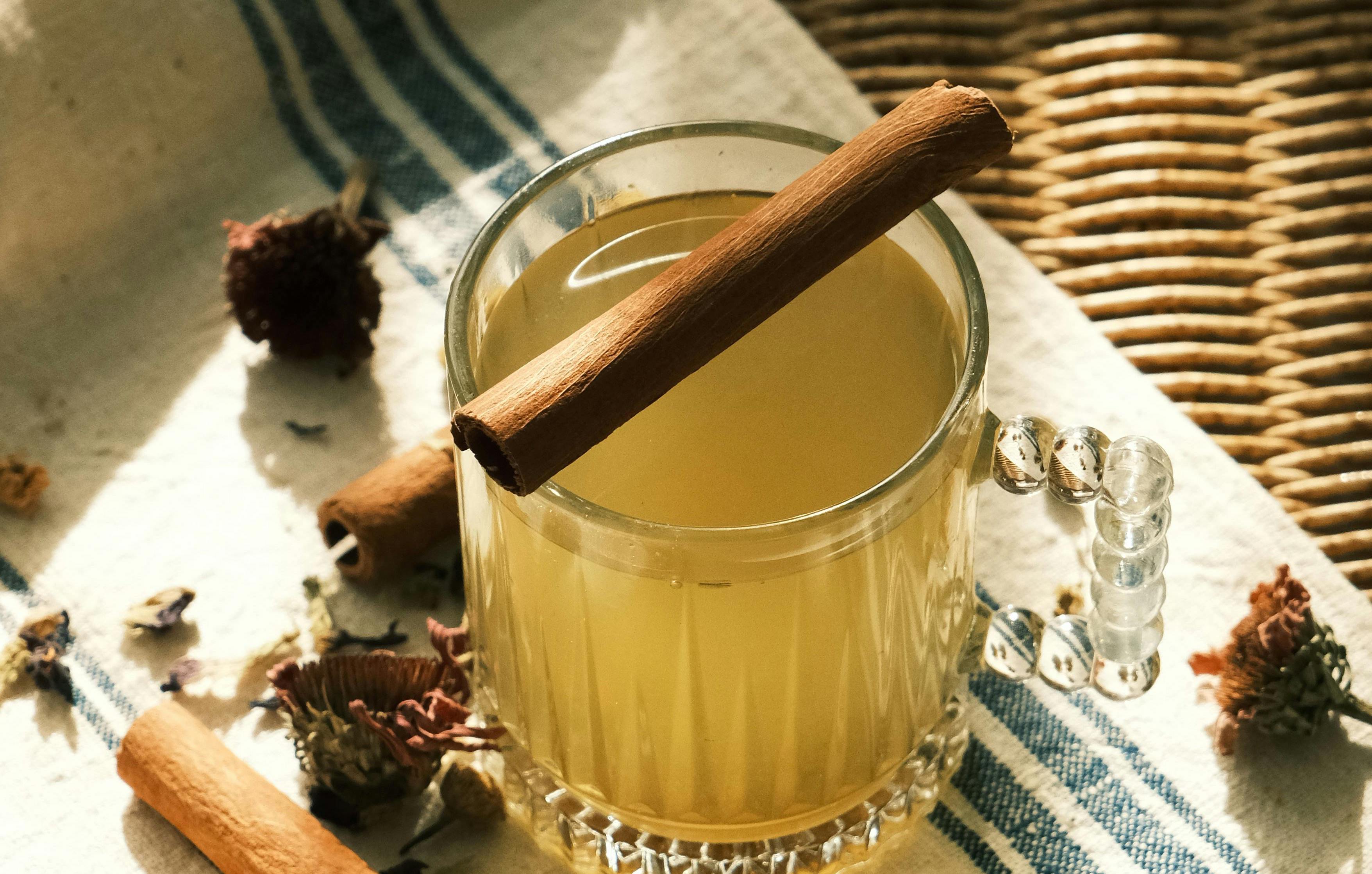 A warm golden drink in a glass mug with a cinnamon stick, surrounded by dried flowers in soft morning light
