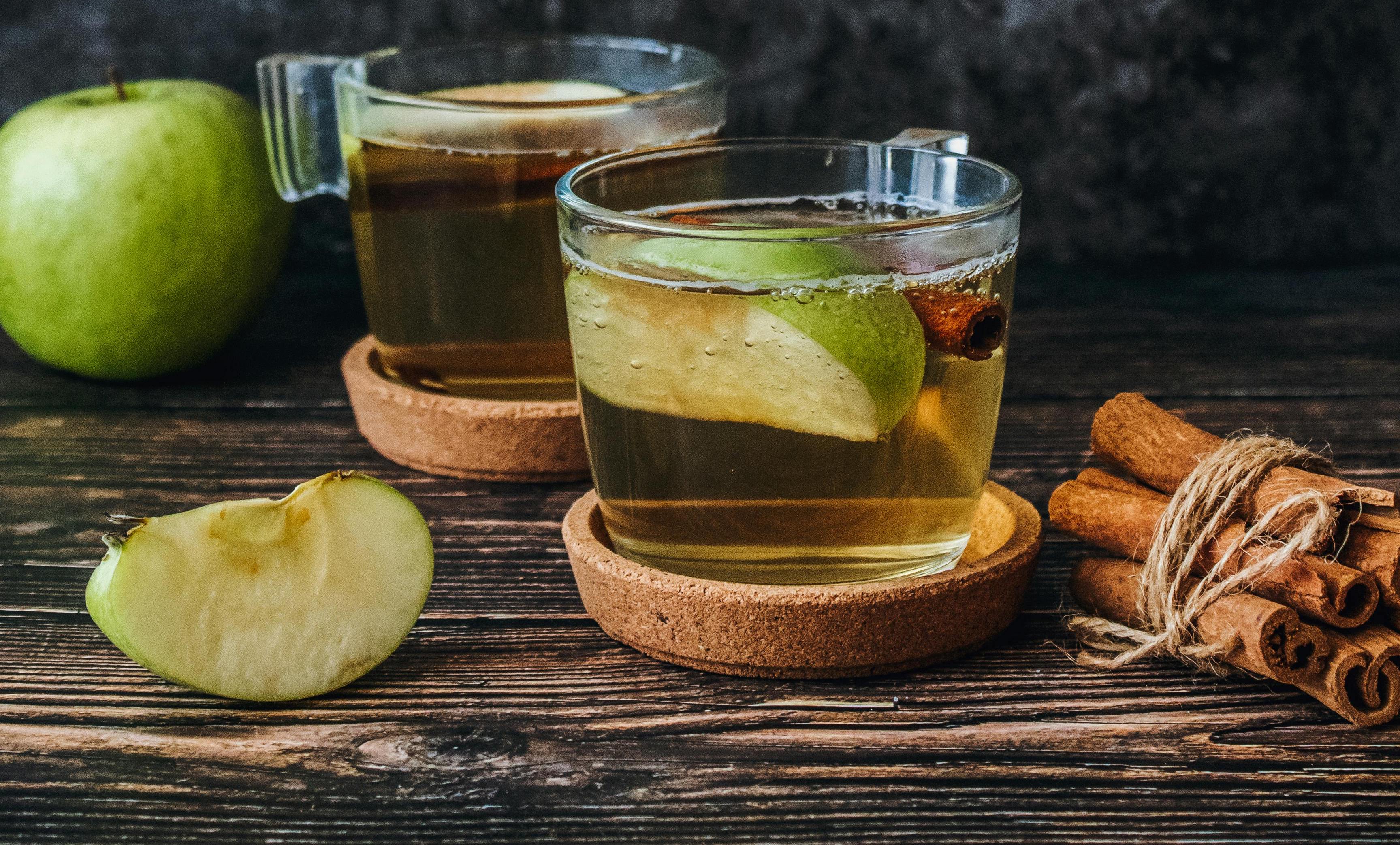 Two glasses of apple cider vinegar drink with green apple slices and cinnamon sticks on a dark wood surface